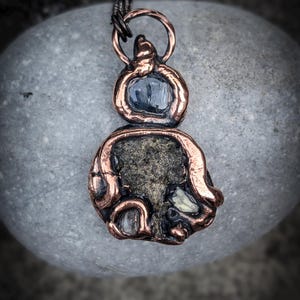May include: A pendant featuring a copper wire-wrapped design with a dark blue stone at the top and a larger, textured, dark metallic stone at the bottom, accented with smaller clear crystals. The text The Magickal Hud Shop is at the bottom.