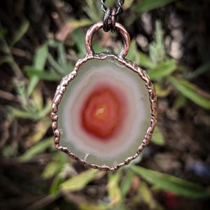 Può includere: Un ciondolo con una fetta di agata circolare traslucida con anelli concentrici bianchi e un centro arancione-rosso vibrante. La pietra è incastonata in una cornice di metallo color rame scuro testurizzato con un occhiello abbinato, appeso a una catena scura.