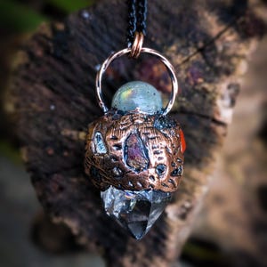 May include: A handcrafted pendant necklace featuring a clear quartz crystal point, a labradorite sphere, and a copper-colored metal setting. The pendant hangs from a black chain. The Magical Hud Shop is printed at the bottom.
