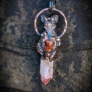 May include: A handcrafted pendant necklace featuring a silver-toned metal charm of a stylized animal figure holding a red-brown stone and a clear quartz crystal. The pendant hangs from a dark chain and is suspended within a copper-colored ring. The Magical Hud Shop.