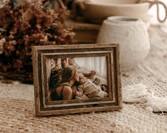 May include: A rectangular picture frame crafted from weathered, natural brown wood with a multi-layered, stepped border design. The frame holds a photograph depicting a person leaning in to kiss a baby, both dressed in neutral-toned clothing.