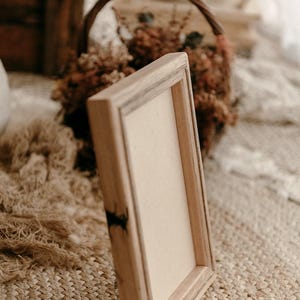 May include: A rustic wooden frame with a blank white canvas inside. The frame is sitting on a woven rug with a basket of dried flowers in the background.