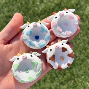 May include: Four handmade cow-themed donut-shaped ornaments. Each ornament is white with colored spots and a cow face design. The colors include blue, pink, green, and brown. The ornaments are held in a hand against a green grass background.