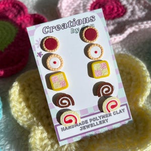 Op de afbeelding: Een set van vijf paar miniatuur oorknopjes in de vorm van eten, gemaakt van polymeerklei en gepresenteerd op een kaart met de tekst Creations by den Handmade Polymer Clay Jewellery. De ontwerpen omvatten taartjes, koekjes en rolletjes.
