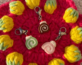Puede incluir: Tres dijes en miniatura con forma de pastel enrollado y cierres de langosta en tono plateado. Los dijes presentan un espiral de chocolate marrón, uno de matcha verde y uno de fresa rosa, cada uno rematado con un pequeño copete de crema blanca.