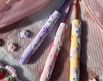 May include: Three crochet hooks with custom handles in lavender, light pink, and pale yellow. Each handle features raised white bunny faces and small bow accents. The hooks are metallic, and the set is accompanied by several small flower charms.