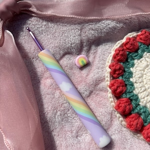Può includere: Un uncinetto con un design arcobaleno e nuvole, un piccolo ciondolo arcobaleno e un sottobicchiere all'uncinetto con accenti floreali rossi. L'uncinetto ha un manico viola e un'estremità ad uncino argentata. Il sottobicchiere ha un centro bianco e un bordo verde.