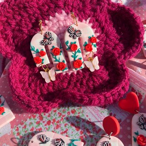 May include: A pair of pink and white arch-shaped earrings with red roses, green leaves, and butterfly accents. The earrings are made of polymer clay and have gold hooks.