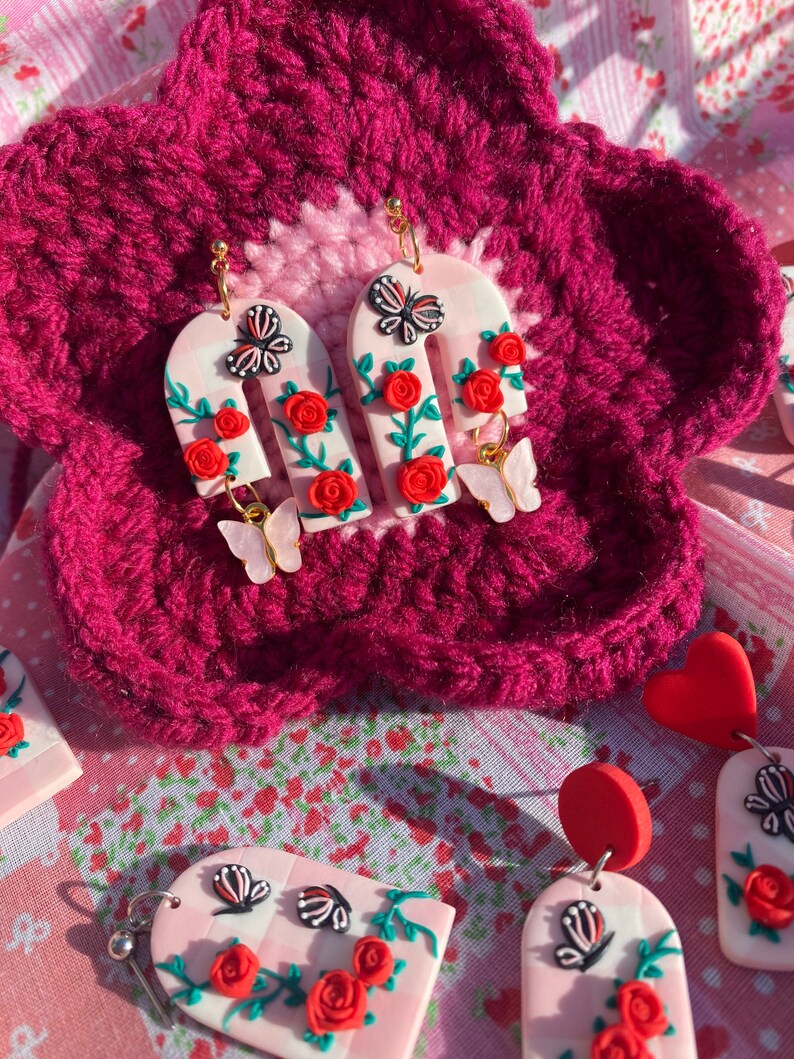 May include: A pair of pink and white arch-shaped earrings with red roses, green leaves, and butterfly accents. The earrings are made of polymer clay and have gold hooks.