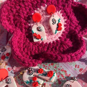 May include: A pair of pink and white arch-shaped earrings with a gingham pattern, featuring red roses and black and white butterflies. The earrings are on a pink and white crocheted surface.