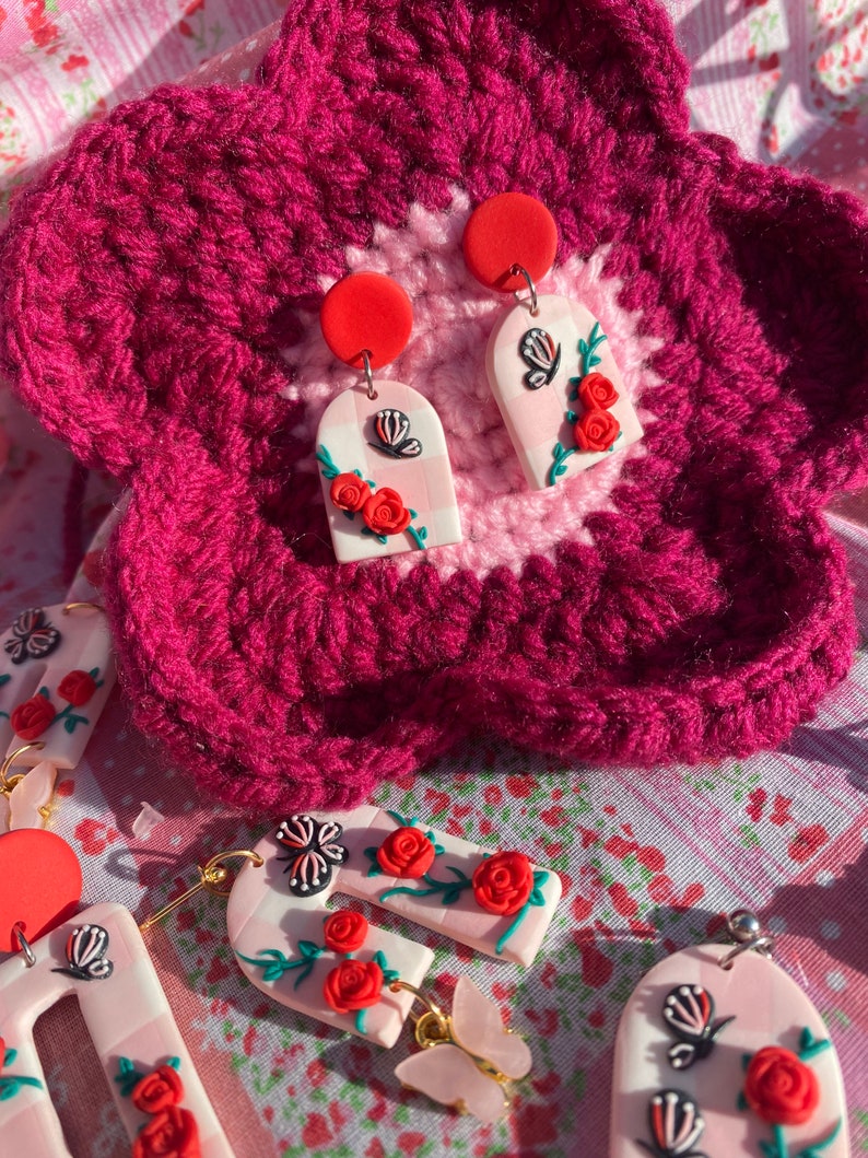 May include: A pair of pink and white arch-shaped earrings with a gingham pattern, featuring red roses and black and white butterflies. The earrings are on a pink and white crocheted surface.