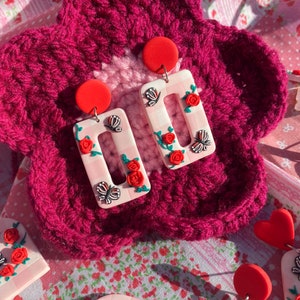 May include: A pair of pink and white rectangular earrings with red roses, green leaves, and black butterflies. The earrings are on a pink and white crocheted background.