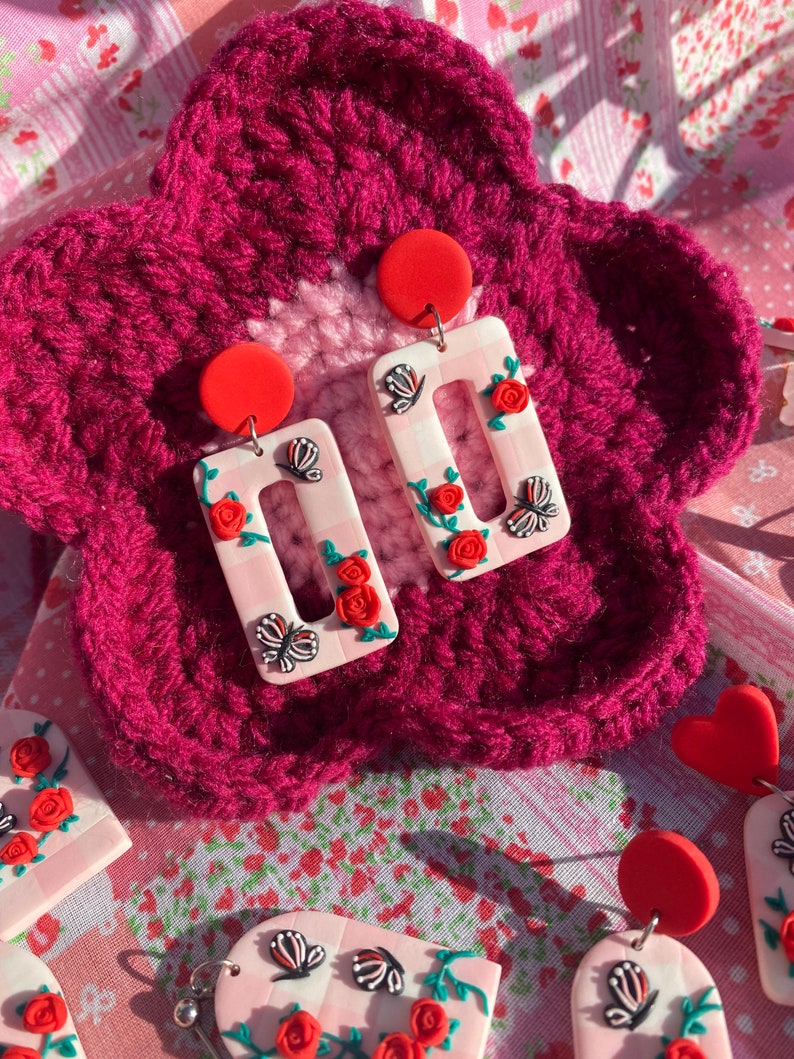 May include: A pair of pink and white rectangular earrings with red roses, green leaves, and black butterflies. The earrings are on a pink and white crocheted background.
