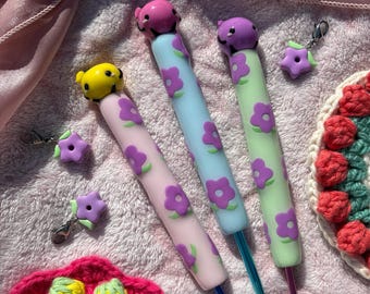 Pode incluir: Três agulhas de crochê com cabos ergonômicos em rosa claro, azul claro e verde claro, decoradas com padrões de flores roxas e finalizadas com uma pequena figura de animal. Três pequenos pingentes de flores roxas com fechos estão incluídos.