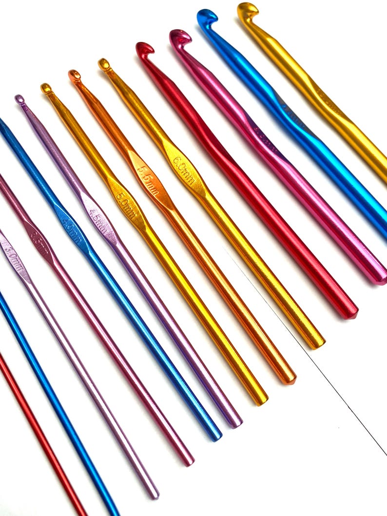 Op de afbeelding: Een verzameling kleurrijke haaknaalden in verschillende maten. De haken zijn gemaakt van metaal en hebben een gladde, glanzende afwerking. De kleuren zijn rood, blauw, goud, paars en roze. De maten staan in millimeters aangegeven.
