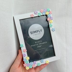 May include: White photo frame decorated with pastel-colored flower embellishments. The frame is labeled "SIMPLY EVERYDAY" and is designed for 4x6 inch photos. Includes easel and hooks for portrait or landscape display.