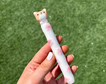 Pode incluir: Uma agulha de crochê branca com detalhes florais rosa e uma alça em forma de gato. O gato tem um rosto marrom e branco com uma gravata borboleta branca. A agulha é segurada em um fundo verde.