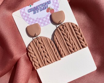 Pode incluir: Um par de brincos de argila marrom em forma de suéter de tricô. Os brincos estão em um cartão branco com o texto "CREATIONS BY DAN".