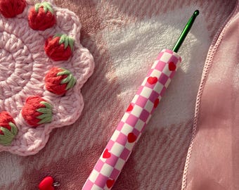Pode incluir: Uma agulha de crochê com um cabo xadrez rosa e branco adornado com corações vermelhos. A agulha tem uma ponta de metal verde. Uma base de morango de crochê rosa e pingentes em forma de coração também são visíveis.