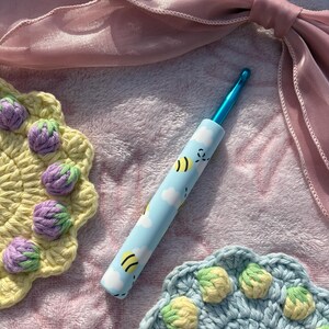 May include: A blue crochet hook with a bee and cloud design, next to two handmade crochet coasters. One coaster is yellow with purple accents, and the other is blue with yellow and green accents. A pink ribbon is in the background.