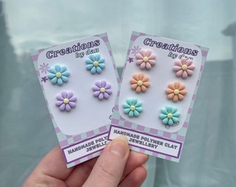 Pode incluir: Dois cartões com um padrão xadrez, cada um exibindo seis brincos de flores de argila polimérica em diferentes cores pastel. Os cartões dizem "Creations by dan" e "Joias de argila polimérica feitas à mão".