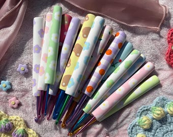 Pode incluir: Uma coleção de agulhas de crochê com cabos estampados apresentando vários designs, incluindo flores, frutas e cores pastel sólidas. As agulhas possuem pontas metálicas em tons de azul, roxo e dourado, dispostas em leque sobre uma superfície macia.