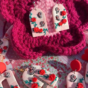 May include: A pair of pink and white arch-shaped earrings with red roses, green vines, and black butterflies. The earrings are made of polymer clay and have silver hooks.