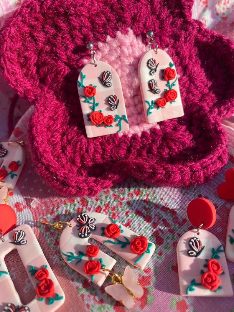 May include: A pair of pink and white arch-shaped earrings with red roses, green vines, and black butterflies. The earrings are made of polymer clay and have silver hooks.