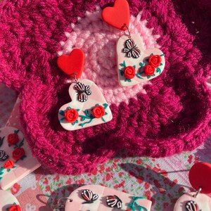 May include: A pair of pink heart-shaped earrings with red hearts at the top, featuring a butterfly and red roses on a vine. The earrings are on a pink and red crocheted surface.