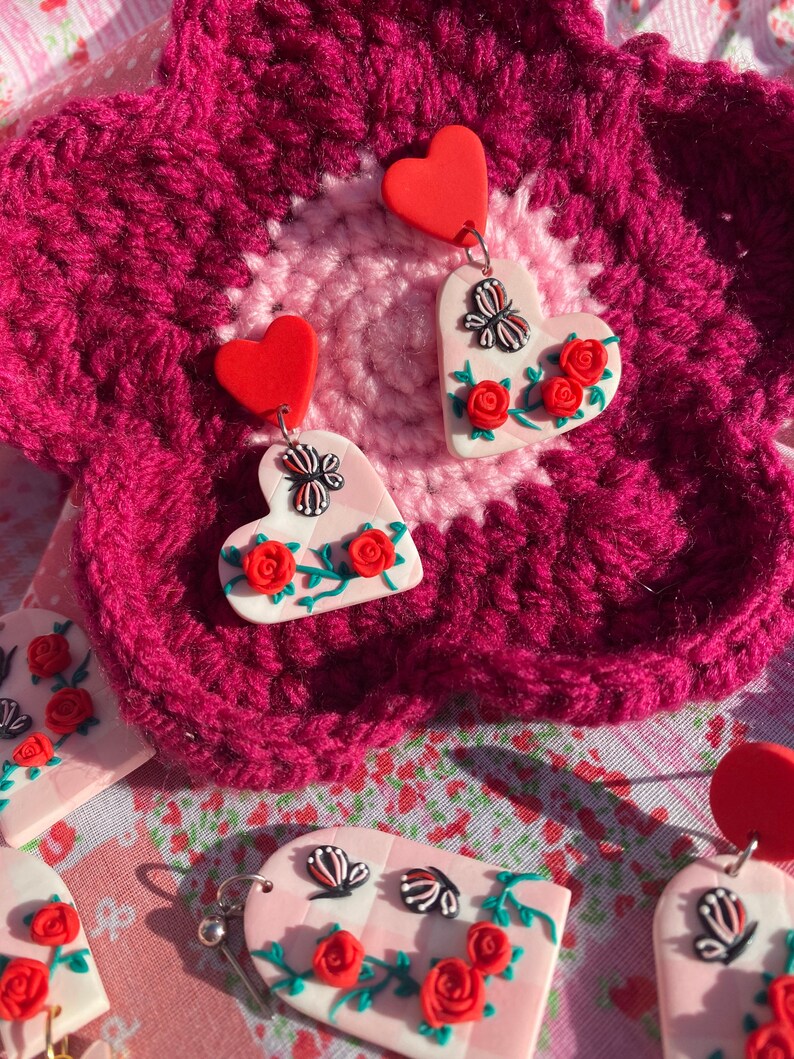 May include: A pair of pink heart-shaped earrings with red hearts at the top, featuring a butterfly and red roses on a vine. The earrings are on a pink and red crocheted surface.