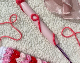Op de afbeelding: Een roze haaknaald met een rood wervelontwerp en een afgeronde bovenkant. De haaknaald ligt naast een stuk roze en rood garen en een gehaakt stuk met rood, roze en wit garen.