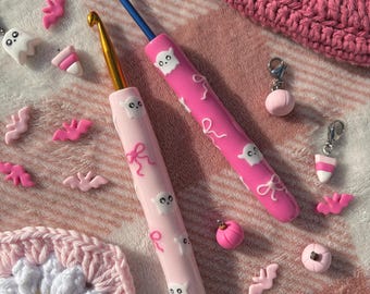 Op de afbeelding: Twee roze en witte haaknaalden met spook- en strikdesigns. De haaknaalden liggen op een roze en witte gehaakte deken met andere roze en witte haakaccessoires.
