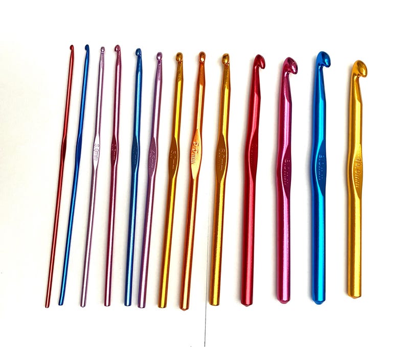 Op de afbeelding: Een verzameling kleurrijke haaknaalden in verschillende maten. De haaknaalden zijn gemaakt van metaal en zijn verkrijgbaar in verschillende kleuren, waaronder rood, blauw, paars, goud en oranje. De naalden liggen op een witte ondergrond.