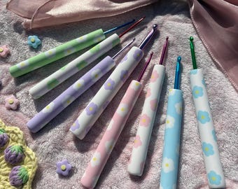Pode incluir: Uma coleção de sete agulhas de crochê com cabos brancos apresentando padrões de flores coloridas em tons de verde, roxo, rosa e azul. Cada agulha tem uma ponta metálica que combina com a cor das flores no cabo, dispostas em formato de leque.