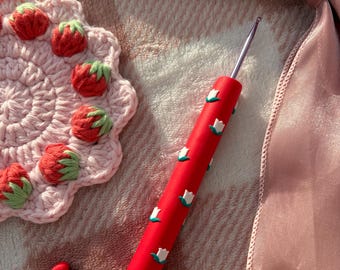 Pode incluir: Uma agulha de crochê vermelha com detalhes de flores brancas e uma ponta de metal prateado repousa sobre uma superfície rosa texturizada. Uma base de crochê rosa em forma de morango e dois pingentes em forma de coração também são visíveis.