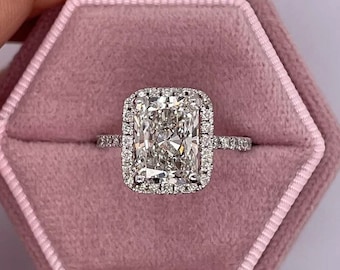 Può includere: Un anello di fidanzamento con diamante di forma quadrata con una montatura a halo. L'anello è incastonato su una fascia in oro bianco ed è presentato in una scatola per anelli in velluto rosa.