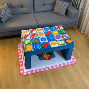 Op de afbeelding: Een blauwe salontafel met een kleurrijk snoep-thema ontwerp, geplaatst op een roze en wit gestreept tapijt. De tafel staat voor een grijze bank met twee zwart-wit geblokte kussens. De kamer heeft houten vloeren.