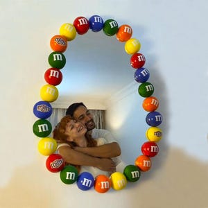 May include: Oval-shaped mirror framed with colorful M&M's candies. The candies are in various colors, including red, yellow, blue, green, and orange. The mirror reflects a couple embracing, creating a playful and vibrant decorative piece.