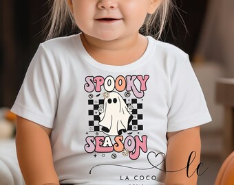 Puede incluir: Una camiseta blanca con un gráfico rosa y naranja de un fantasma con el texto "Spooky Season" en una fuente retro. El fantasma sostiene signos de paz y tiene una flor en la cabeza. El gráfico está sobre un fondo a cuadros.