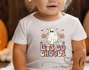 Puede incluir: Una camiseta blanca con un lindo fantasma que lleva una corona de flores y hace el signo de la paz. La camiseta tiene el texto "Let's Go Ghouls" en letras rosas y naranjas.