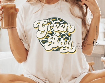 Puede incluir: Camiseta blanca con un gráfico verde y amarillo que dice "Green Bay" en una fuente retro. El gráfico está rodeado por un patrón de estampado de leopardo.