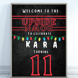 May include: Framed black poster with red and white text. The text reads "WELCOME TO THE UPSIDE NMOG TO CELEBRATE KARA TURNING 11". A string of colourful lights is at the bottom. The poster is in a wooden frame.