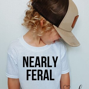 May include: A white t-shirt with the text "NEARLY FERAL" printed in black. The shirt is being worn by a child wearing a brown and black mesh baseball cap. The text "LA COCO DESIGN" is printed in black at the bottom of the shirt.