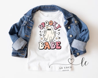 Puede incluir: Camiseta blanca con un gráfico de "Spooky Babe" en rosa, melocotón y negro. El diseño presenta un fantasma haciendo el signo de la paz, estrellas y flores. Una chaqueta vaquera está sobre la camiseta.