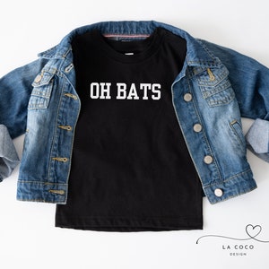 Può includere: Maglietta nera con testo bianco che dice "OH BATS". La maglietta è piegata e appoggiata su una giacca di jeans blu.