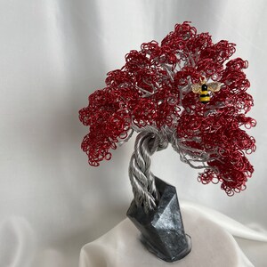 Può includere: Una scultura di un albero in filo di ferro rosso con un piccolo ciondolo a forma di ape dorata su un ramo. L'albero è su una base di pietra grigia.