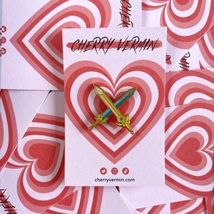 Puede incluir: Pin de esmalte de espada dorada y azul sobre una tarjeta blanca con diseño de corazón rojo. La tarjeta tiene el texto "CHERRY VERMIN" e iconos de redes sociales. El fondo presenta múltiples tarjetas con el mismo diseño de corazón.