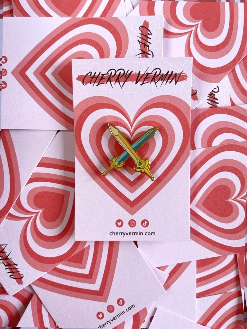 May include: A gold and blue sword enamel pin on a white card with a red heart design. The card has the text "CHERRY VERMIN" and social media icons. The background features multiple cards with the same heart design.