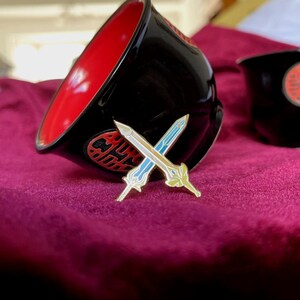 May include: A black bowl with a red interior and a decorative emblem, next to a crossed swords pin with blue and gold accents. The pin is on a purple surface. The bowl has a Chinese character emblem.