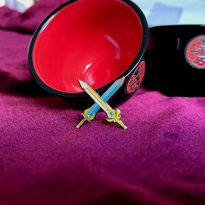 May include: Two crossed sword pins with gold and blue enamel detailing, set against a deep purple background. The pins are in front of a black and red bowl with a decorative design.